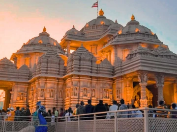 A grand view of Akshardham Temple in Delhi, featuring intricate pink sandstone and white marble carvings, with lush gardens and a serene water body reflecting its majestic structure under a clear sky.