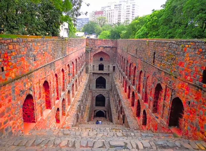 A mesmerizing view of Agrasen Ki Baoli in Delhi, a historic stepwell with deep red sandstone walls and arched niches, descending multiple levels with a few visitors exploring, surrounded by lush greenery and modern buildings in the background.