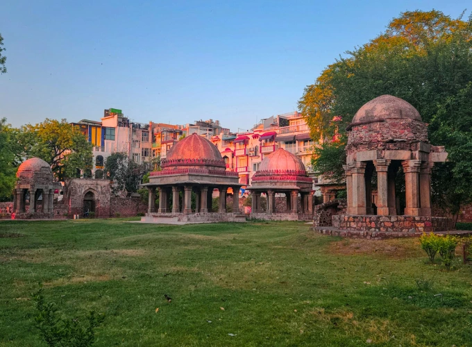 A serene view of Hauz Khas Complex in Delhi, featuring ancient red sandstone tombs with domed roofs surrounded by lush green lawns and a historic water tank, with modern buildings visible in the background under a clear evening sky.