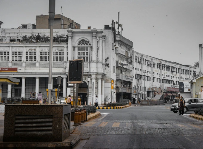 A serene view of Connaught Place in Delhi, featuring elegant colonial-style buildings with arched windows and a few pedestrians, set against a quiet street with modern shops and cars under a cloudy sky.