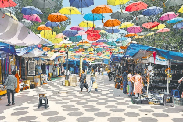 A bustling Delhi Haat market with a canopy of colorful umbrellas overhead, showcasing vibrant stalls filled with goods and people shopping on a patterned walkway.
