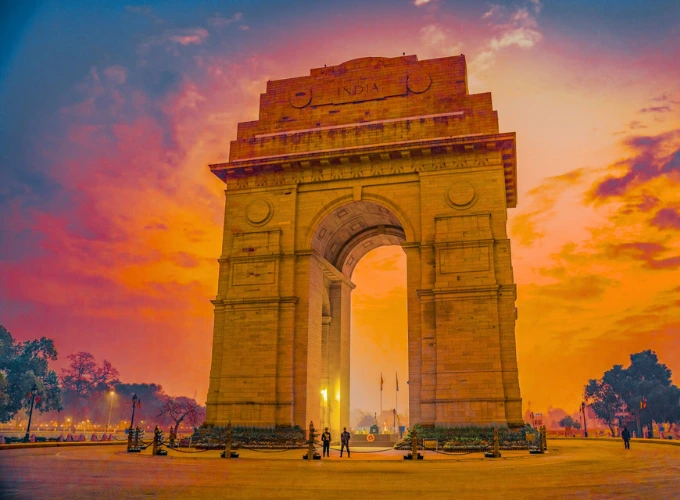 A stunning view of India Gate in Delhi at sunset, with the iconic war memorial archway glowing against a vibrant orange and pink sky. A few people stand near the base, surrounded by a spacious plaza and distant trees.