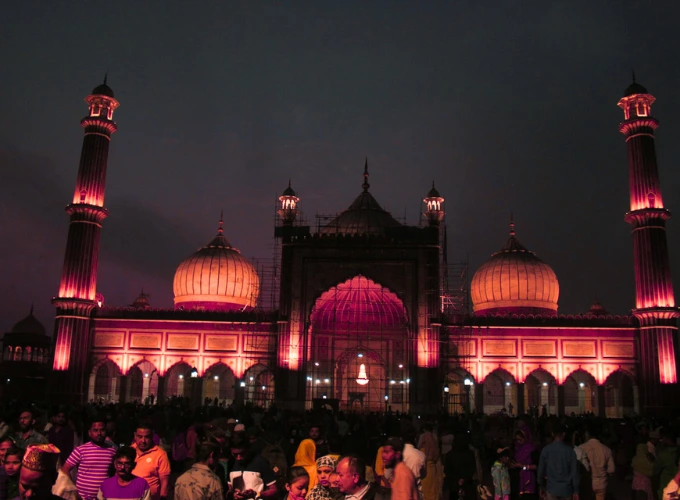 A vibrant night view of Jama Masjid in Delhi, beautifully illuminated with pink and purple lights, showcasing its grand minarets and domes. A crowd of people gathers in the foreground, adding liveliness to the scene.
