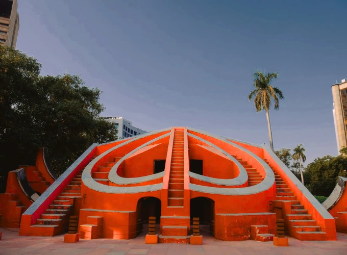 A striking view of Jantar Mantar in Delhi, featuring a vibrant orange astronomical instrument with intricate staircases and arches, set against a clear blue sky with trees and modern buildings in the background.