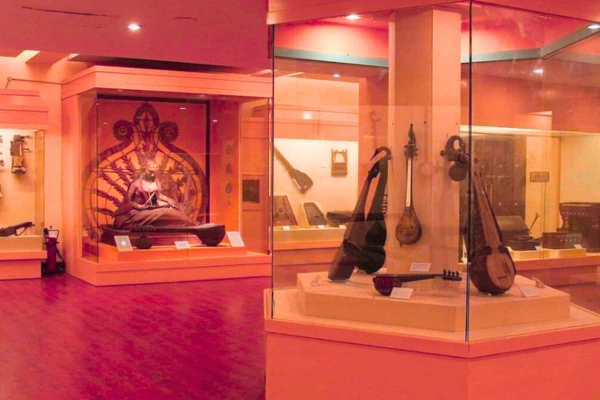 An exhibit in a Delhi museum displaying a large statue of a deity and traditional musical instruments in glass cases, with warm lighting and red flooring.