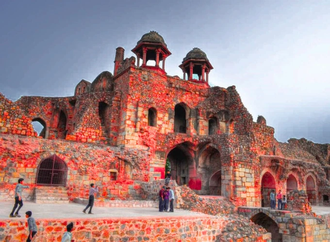 A striking view of Purana Qila in Delhi, showcasing its ancient red sandstone ruins with arched gateways and domed pavilions, bathed in warm light. A few visitors explore the historic fort against a cloudy sky.
