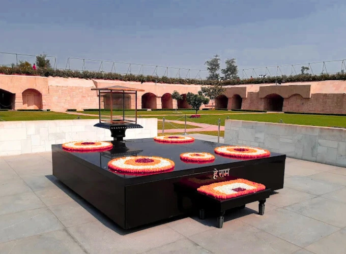 A solemn view of Raj Ghat in Delhi, featuring Mahatma Gandhi's memorial with a black marble platform adorned with vibrant floral wreaths and the eternal flame, set against a backdrop of green lawns and arched walls under a clear sky.